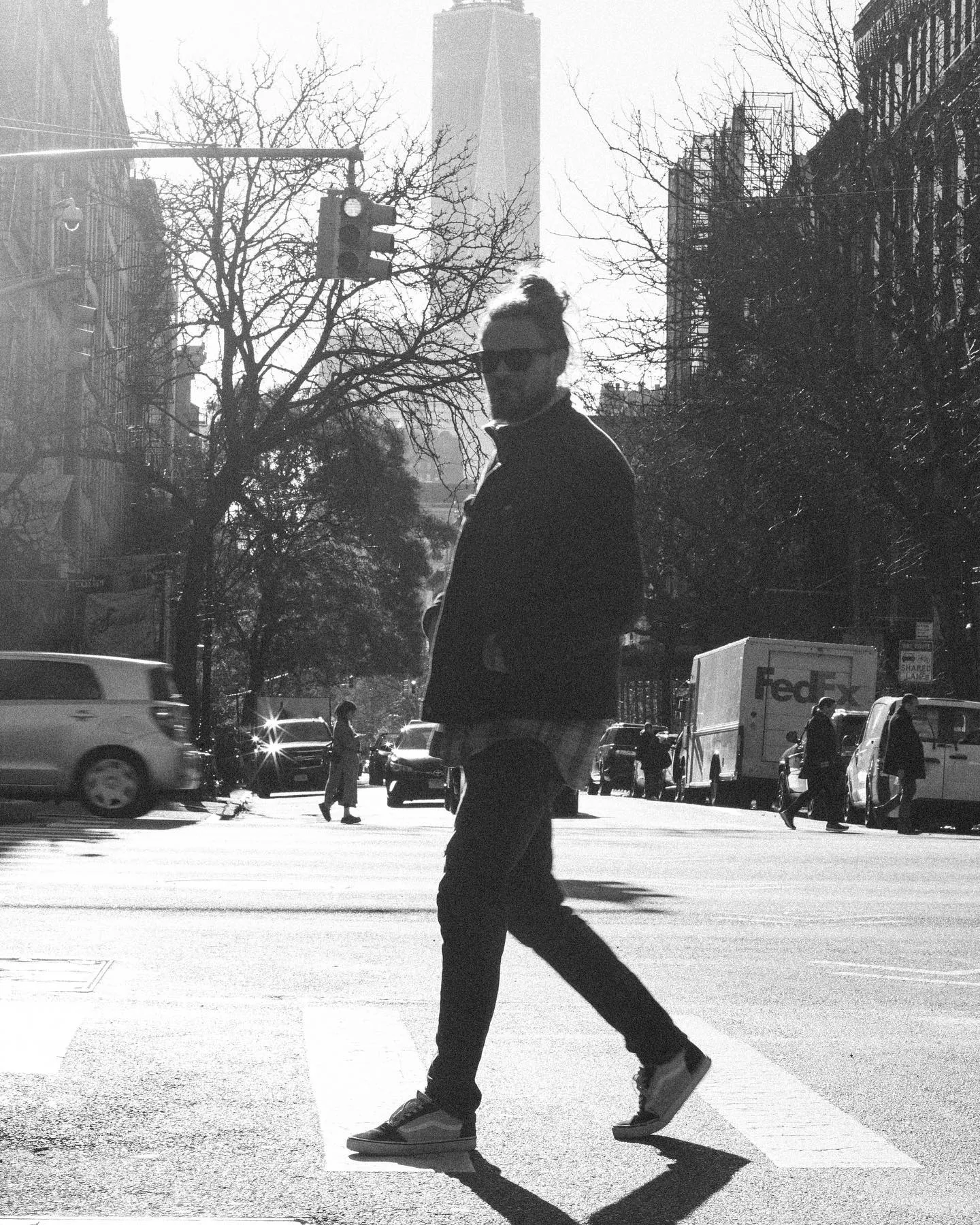 Backlit black-and-white street frame: a man in sunglasses and a dark jacket strides across a painted crosswalk arrow in low winter sun, bare trees, a FedEx truck and the tower of One World Trade rising in the haze behind him.
