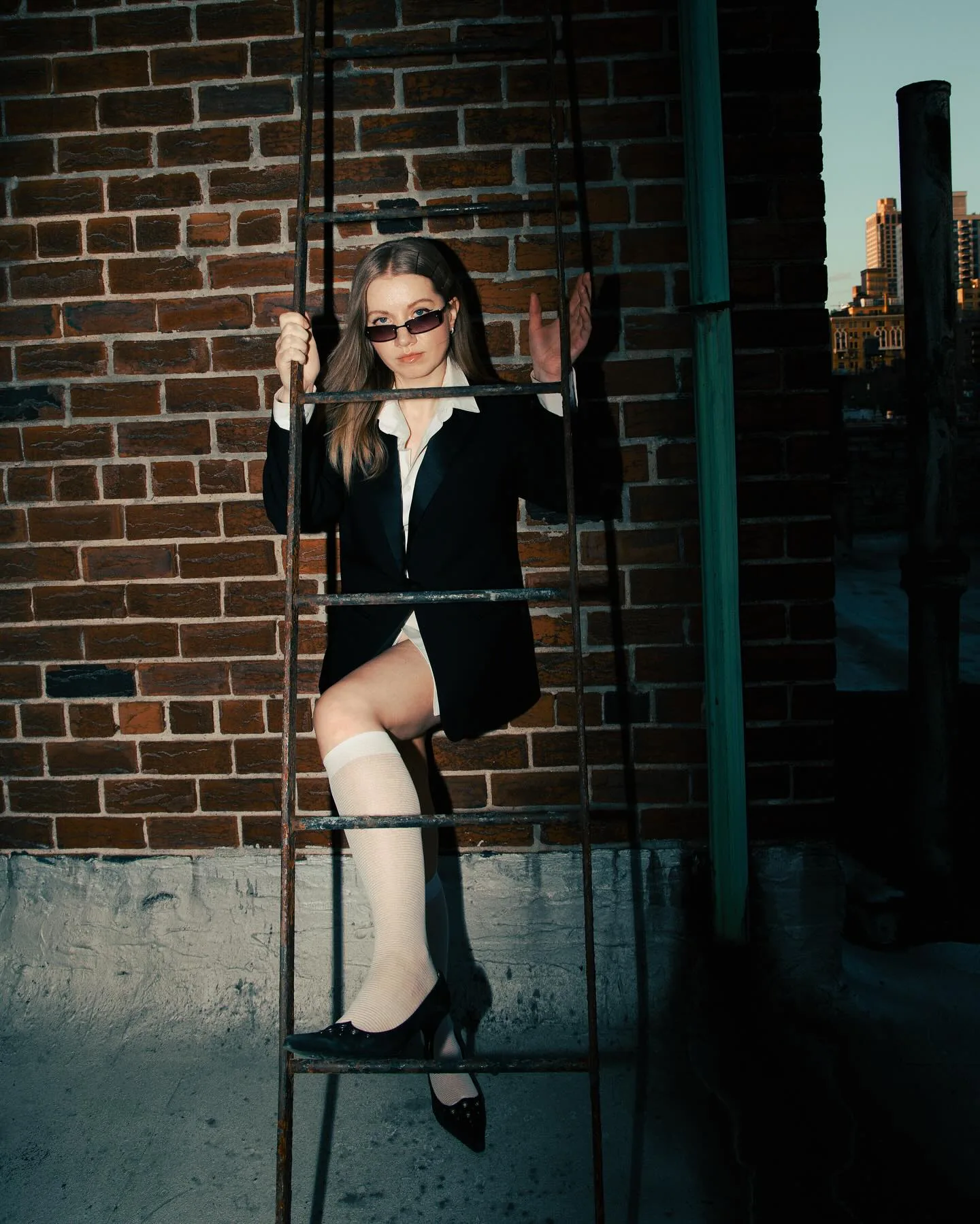 Woman in a black blazer, white shirt, knee socks and heels perched on a rooftop fire escape ladder against red brick, low sun over the Manhattan skyline behind her.