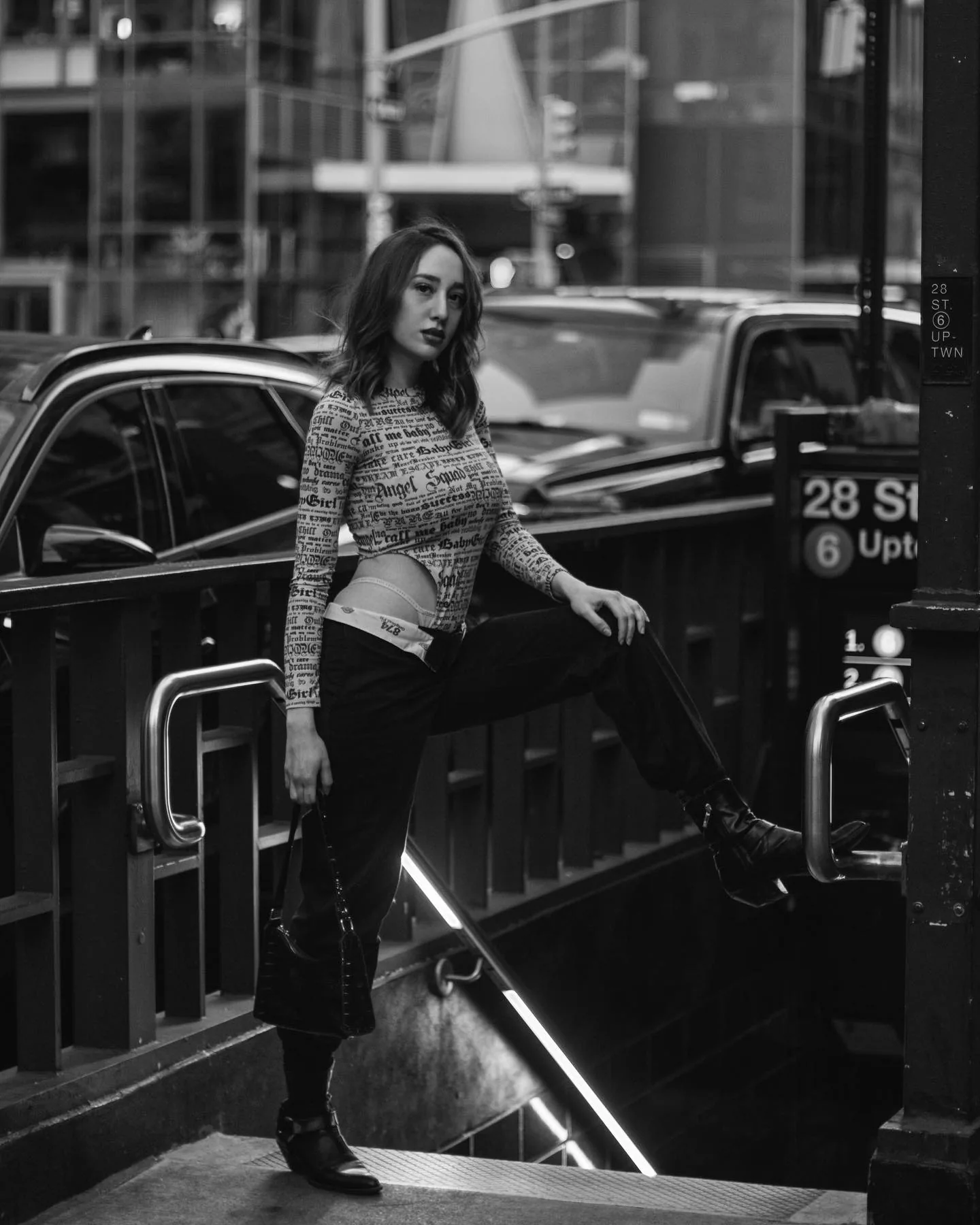Black-and-white frame of a woman in a newsprint-patterned long-sleeve top and black trousers, one boot braced on the subway stair rail at the 28 St 6 Uptown entrance, holding a small black bag, cars and glass buildings behind her.