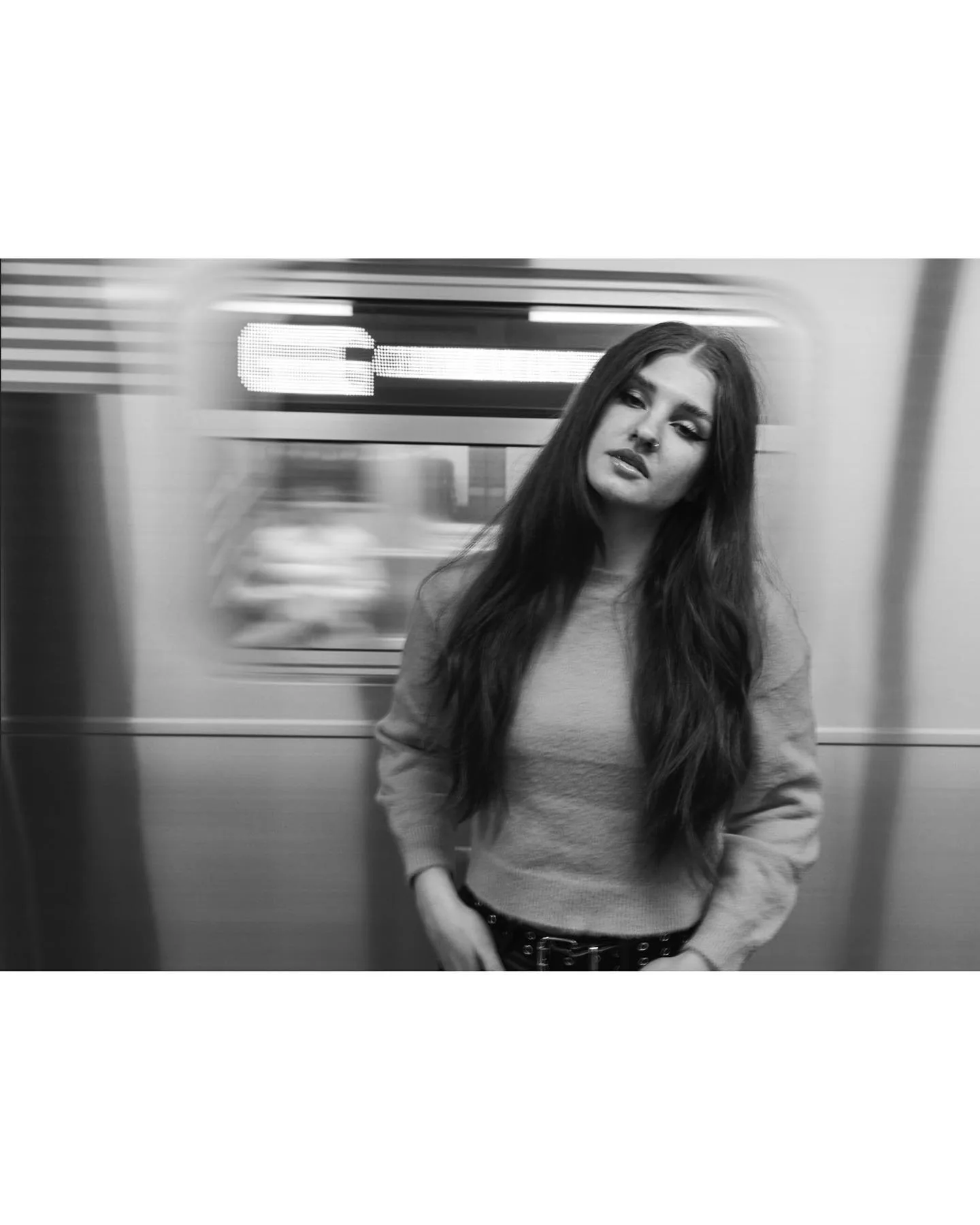 Black-and-white frame of a woman standing still on a subway platform in a pale sweatshirt and studded belt, head tilted, as a train streaks past behind her in heavy motion blur.
