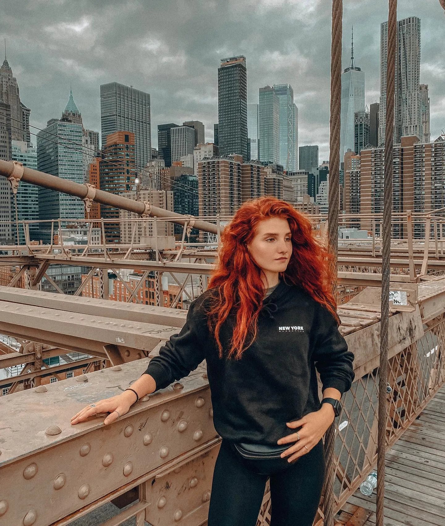 Daria on the Brooklyn Bridge walkway leaning against a riveted steel beam, black NEW YORK MANHATTAN hoodie and black leggings, wind in her bright red hair, the Lower Manhattan skyline and One World Trade under heavy grey cloud behind her.