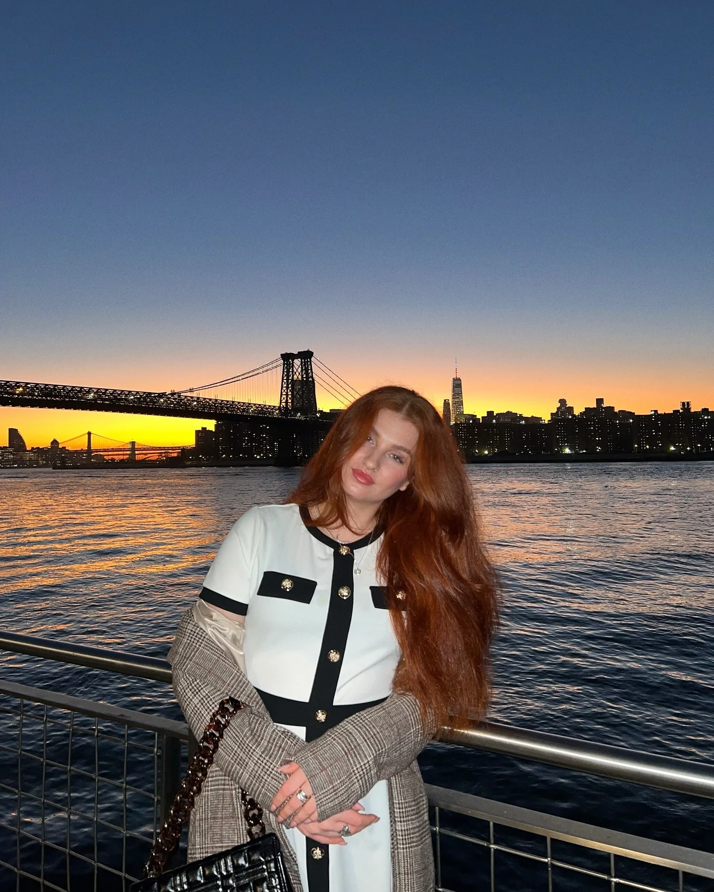 Daria on the Brooklyn waterfront in a black-and-white tweed jacket and plaid trousers, seated against the railing with the Williamsburg Bridge and Manhattan skyline in orange sunset behind her