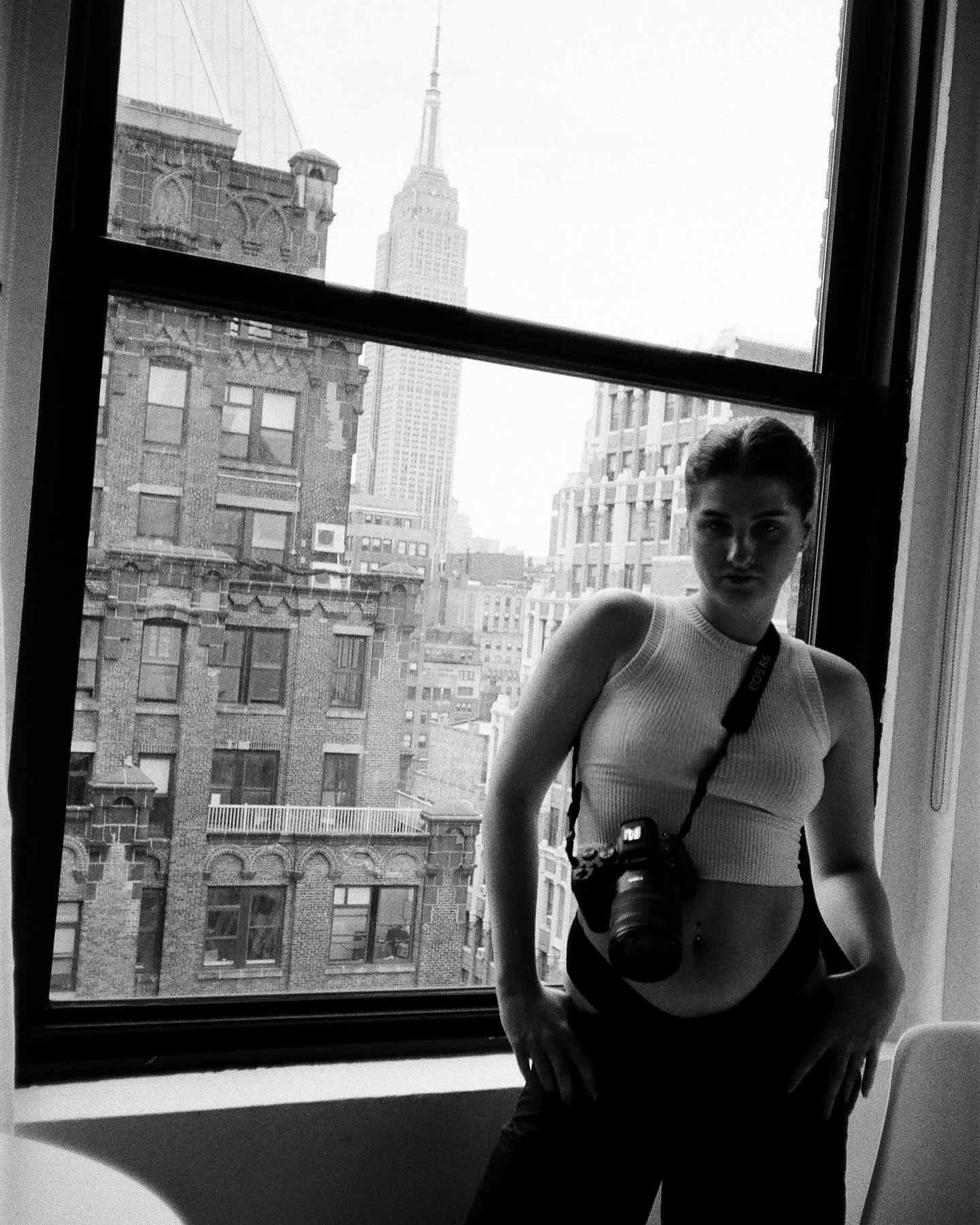 Black-and-white film frame of Daria standing in an open apartment window in a white tank top with a 35mm camera hanging at her hip, the Empire State Building and Manhattan brick facades behind her.
