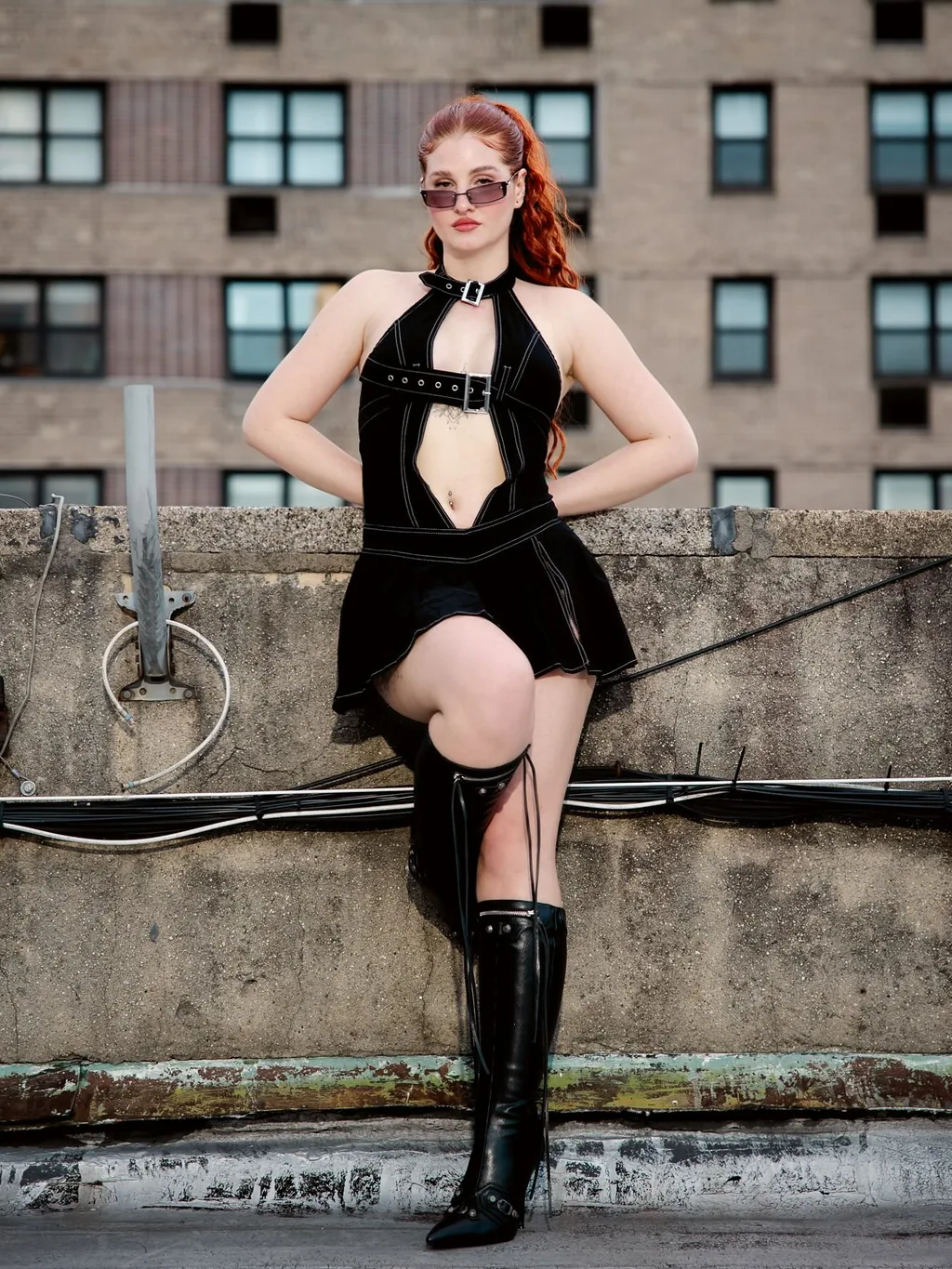 Full-length frame of Daria against a rooftop parapet in a black cutout mini dress and tall black boots, a grid of red brick apartment windows filling the frame behind her