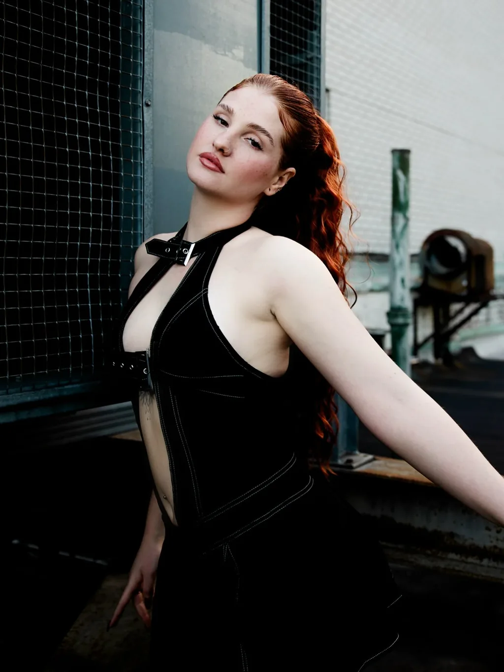 Full-length shot of Daria in a black cut-out dress leaning back against a rooftop chain-link fence, one leg extended, Manhattan water towers and brick facades behind
