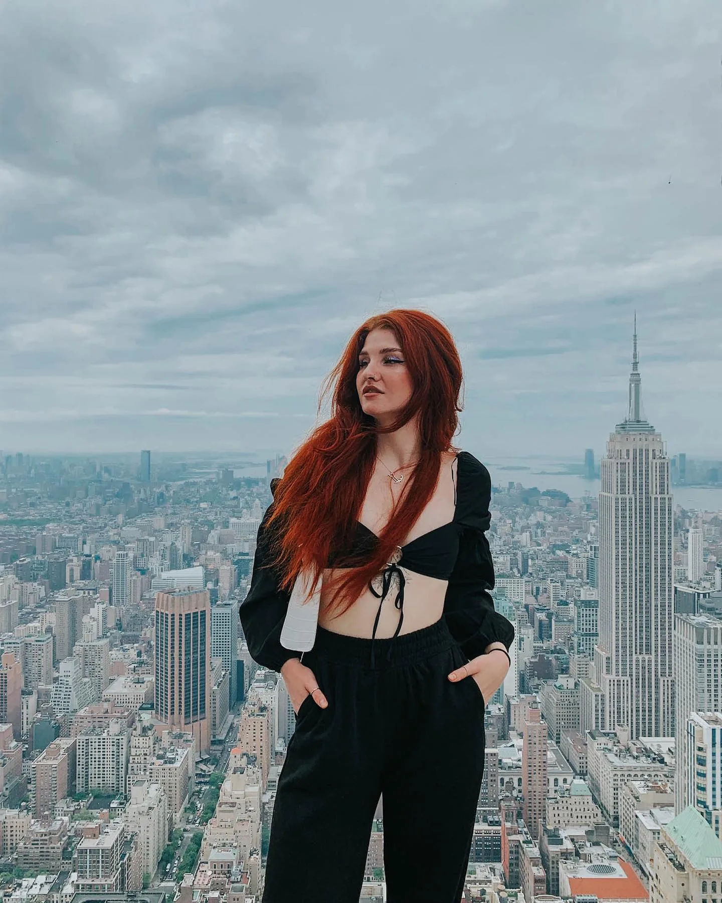 Woman with long red hair in a black puff-sleeve crop top and wide black trousers, hands in pockets, standing high above the Manhattan grid with the Empire State Building rising beside her under a heavy grey sky.