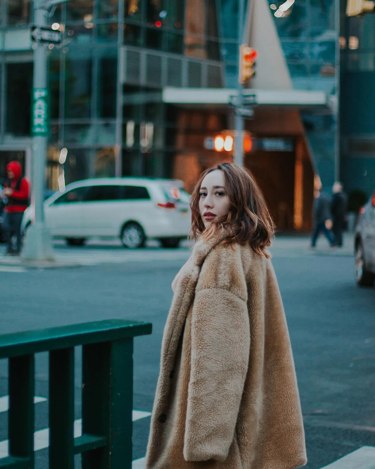 A woman in an oversized camel teddy-fur coat pauses mid-crossing on a Manhattan street at dusk, glancing back over her shoulder; a white minivan, glass tower and traffic light blur behind her in cool blue evening light.