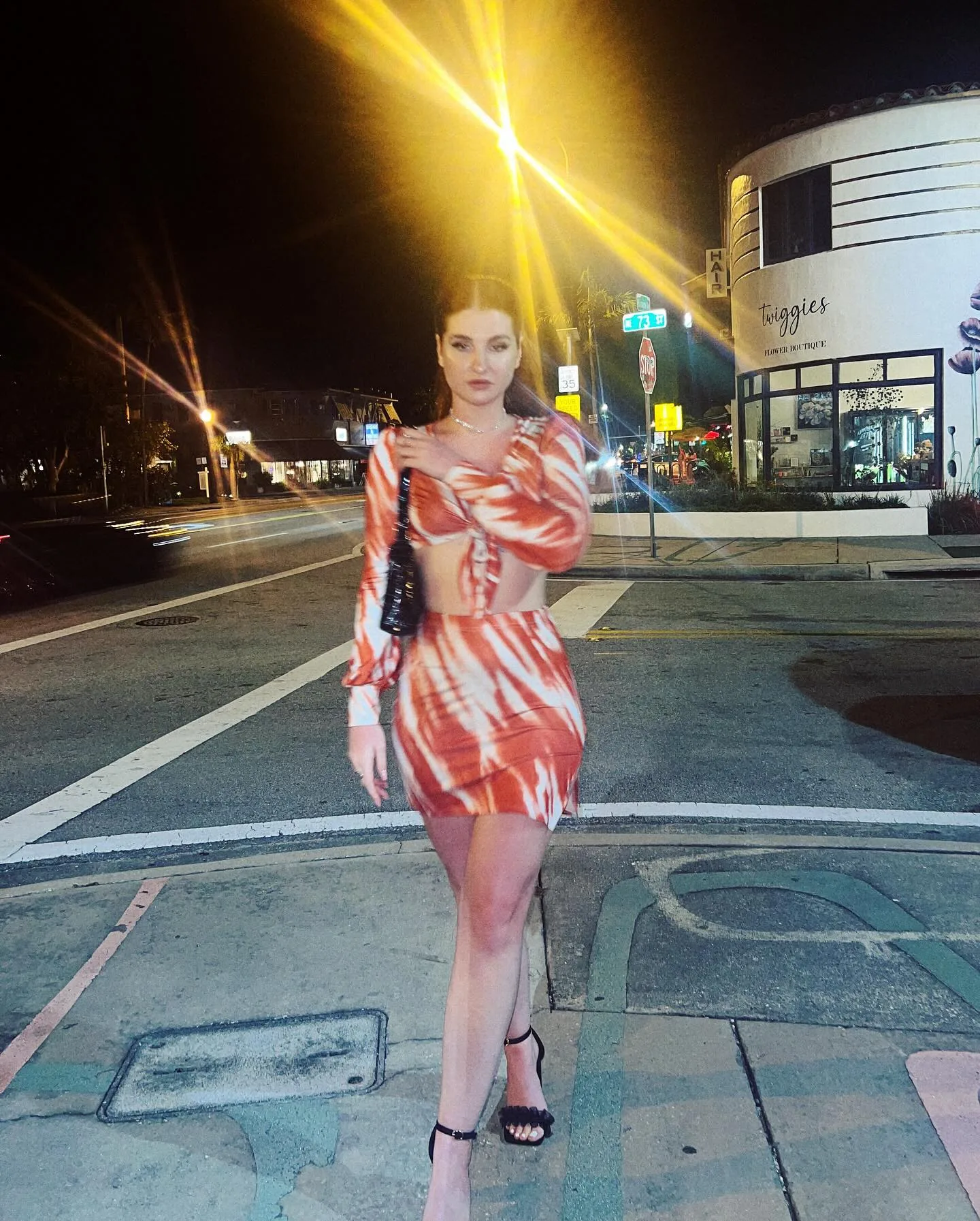 Daria standing alone in the middle of an empty city street at night in a marbled orange-and-white cutout dress and black heels, streetlamp flare above her, shopfronts and road markings receding behind