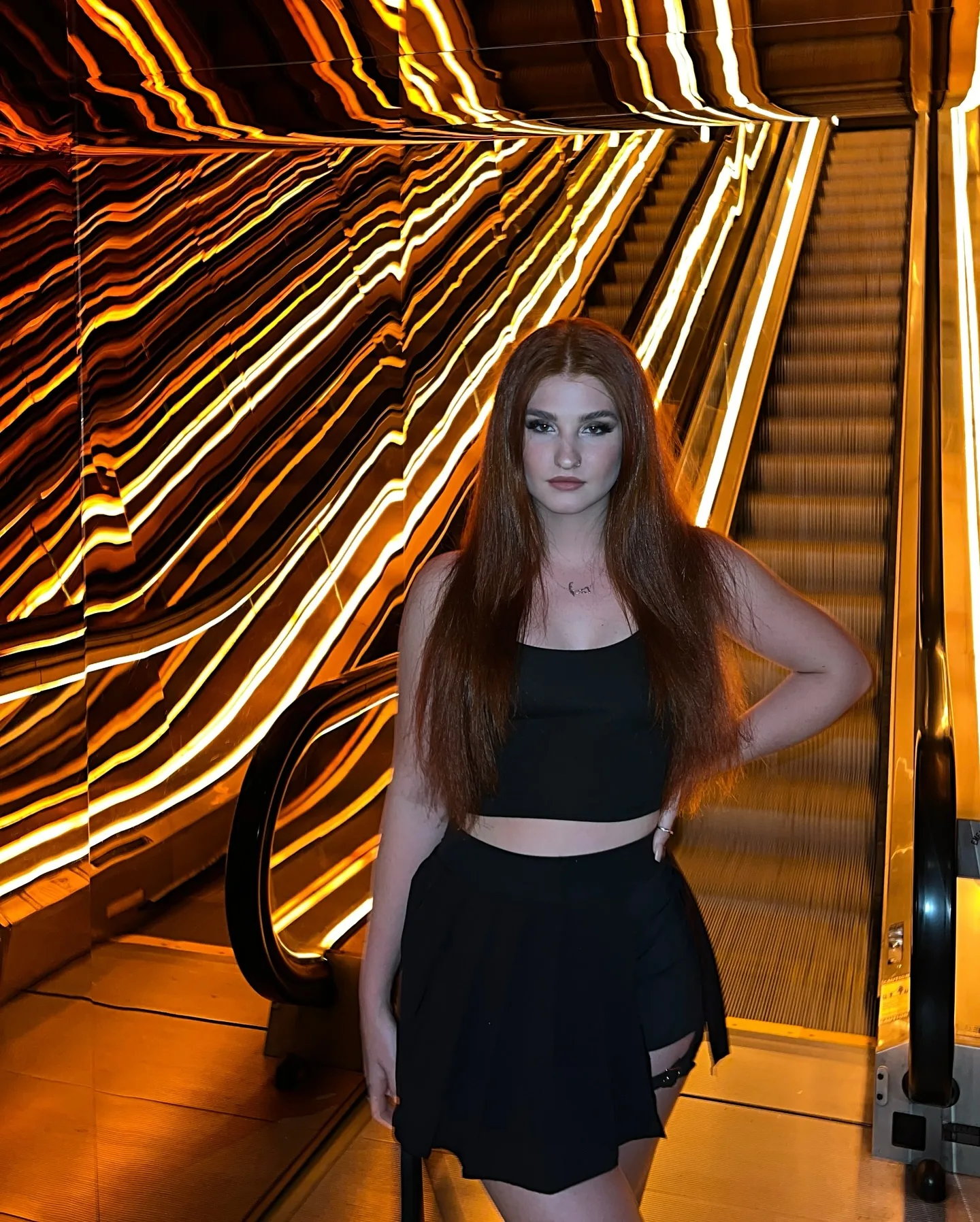 Daria standing at the foot of an escalator in a black crop top and pleated black skirt, long copper hair down, the entire wall beside her exploding into warped ribbons of gold and white light.