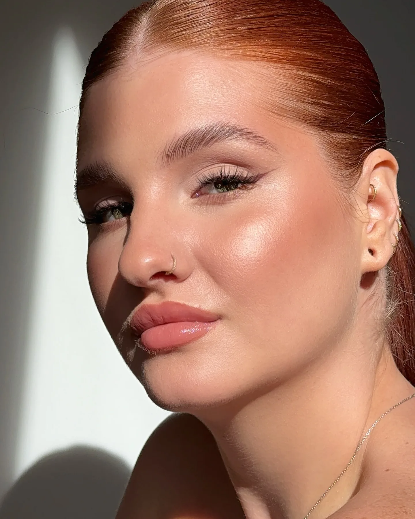 Beauty close-up of Daria filling the frame, copper hair slicked back off her forehead, glossy skin and a small gold nose ring, looking straight into the lens with a hard wedge of sunlight falling on the pale wall behind her