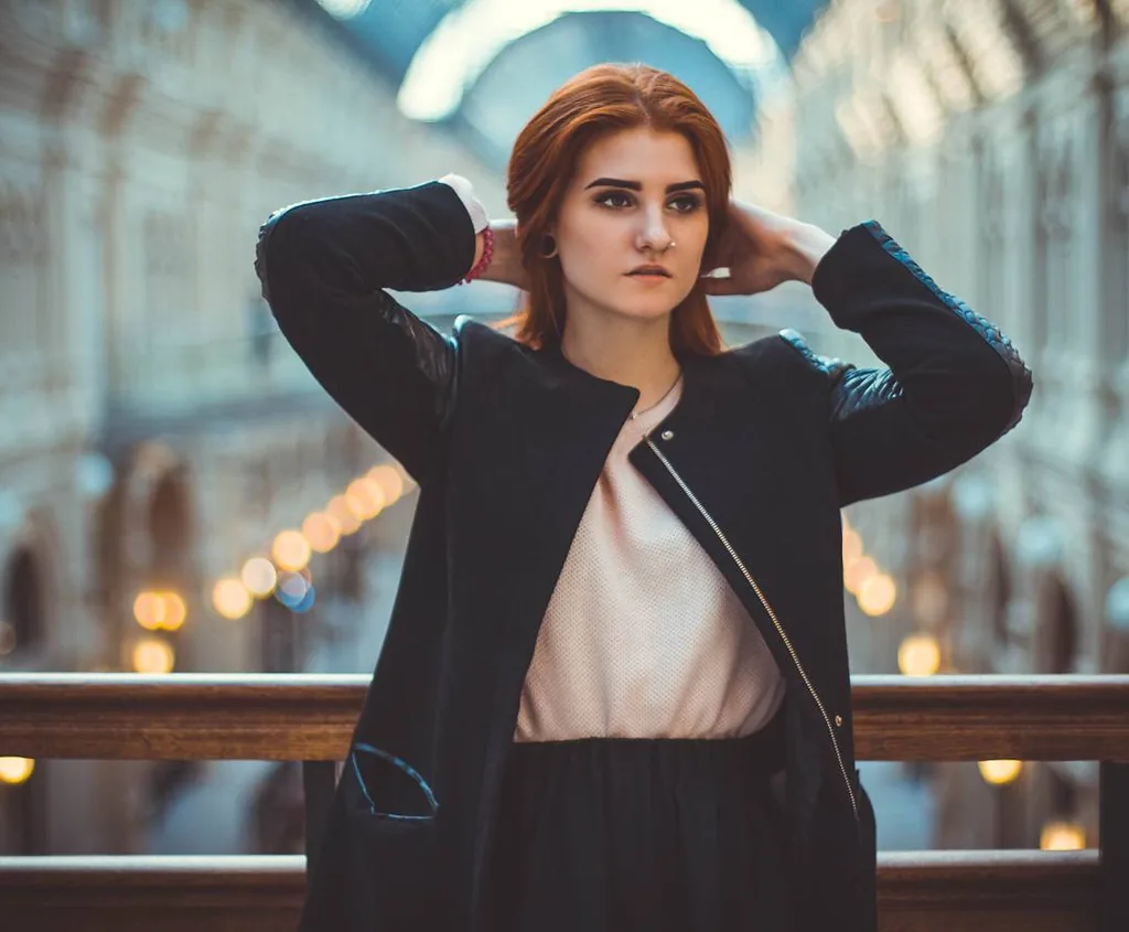 Daria in a black coat over a blush blouse, both hands lifted into her red hair, standing at a wooden balustrade in the glass-roofed GUM arcade with strings of lights receding behind her