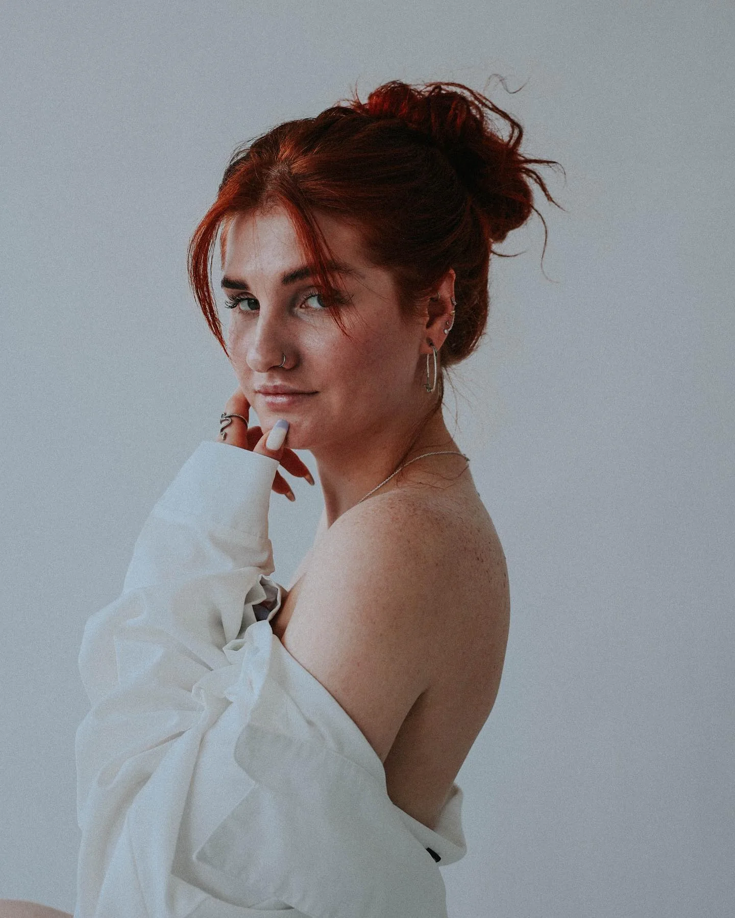Daria looking back over one bare shoulder in a loose white shirt pulled off the shoulder, red hair in a messy topknot, fingers at her chin, against a soft grey-green backdrop.