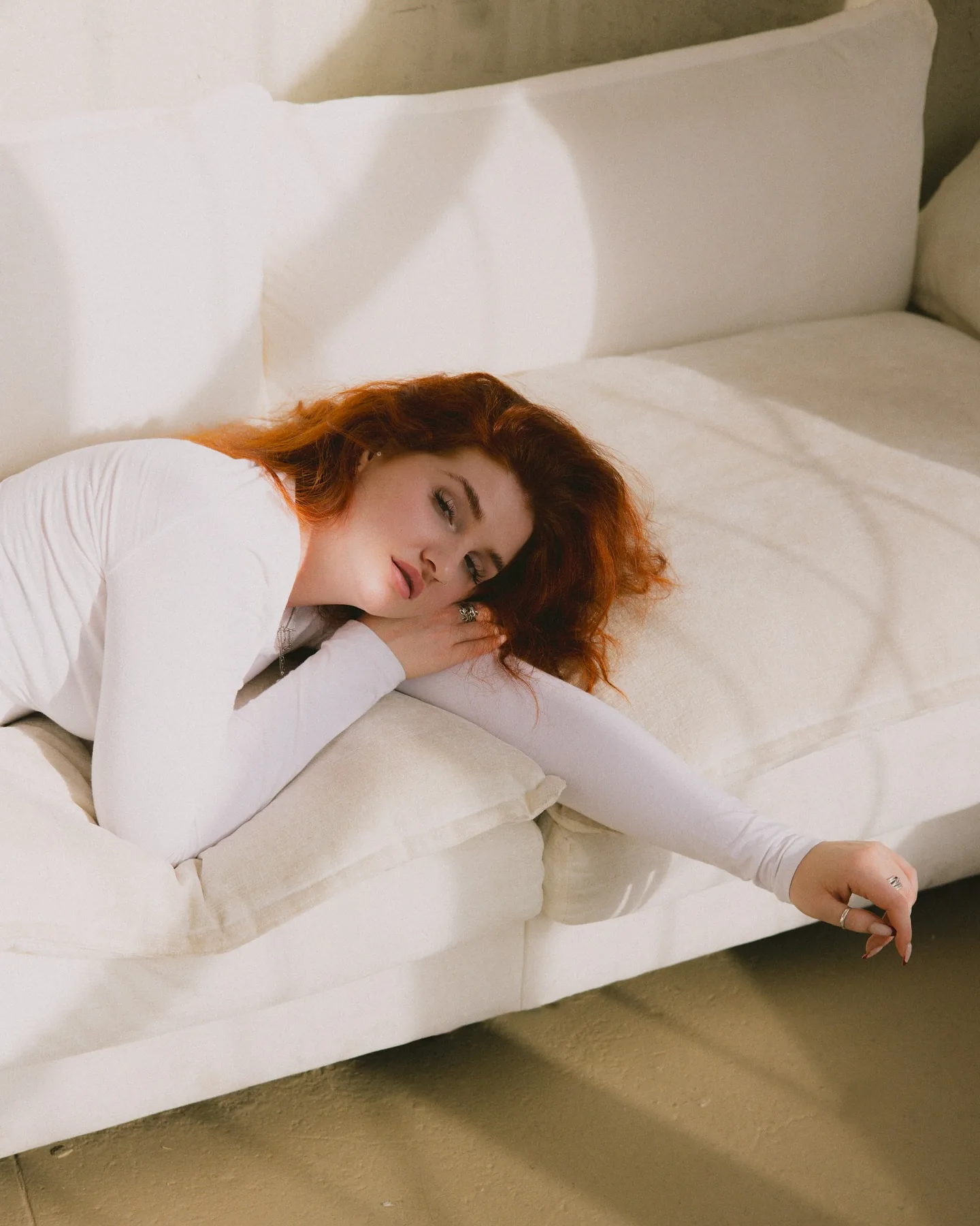 Daria lying across a cream linen sofa in a white long-sleeve top, eyes closed, red hair fanned out over the cushion, soft window light and a faint shadow raking the wall behind