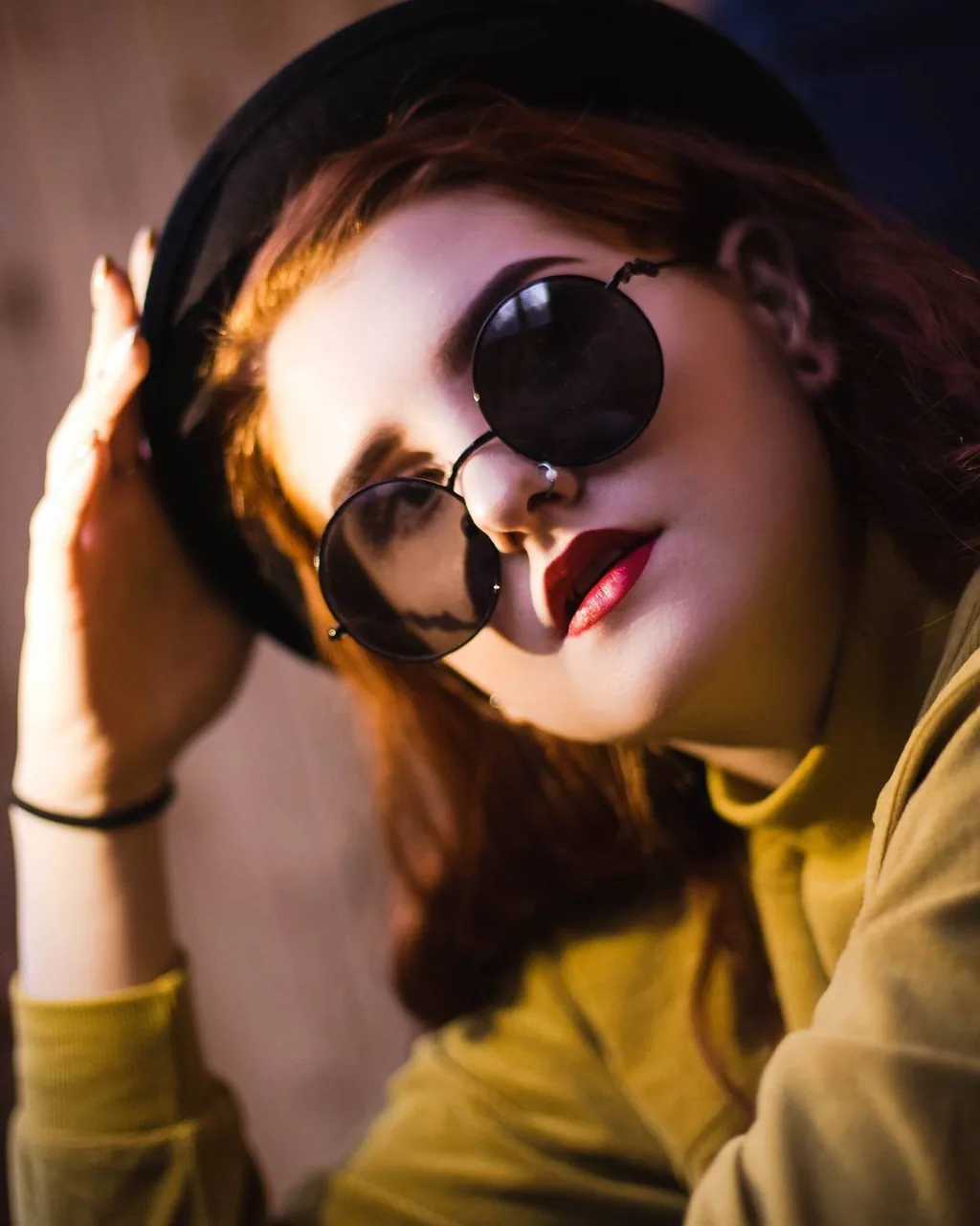 Close portrait of Daria tilted against a wooden wall in a black beret and round black wire-rim sunglasses, chartreuse turtleneck, deep red lipstick, one hand raised to the hat brim.
