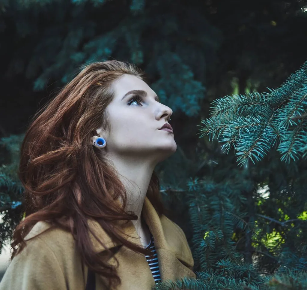Daria in profile in a camel wool coat, chin lifted, red hair blown across her shoulder, against dark blue-green spruce branches