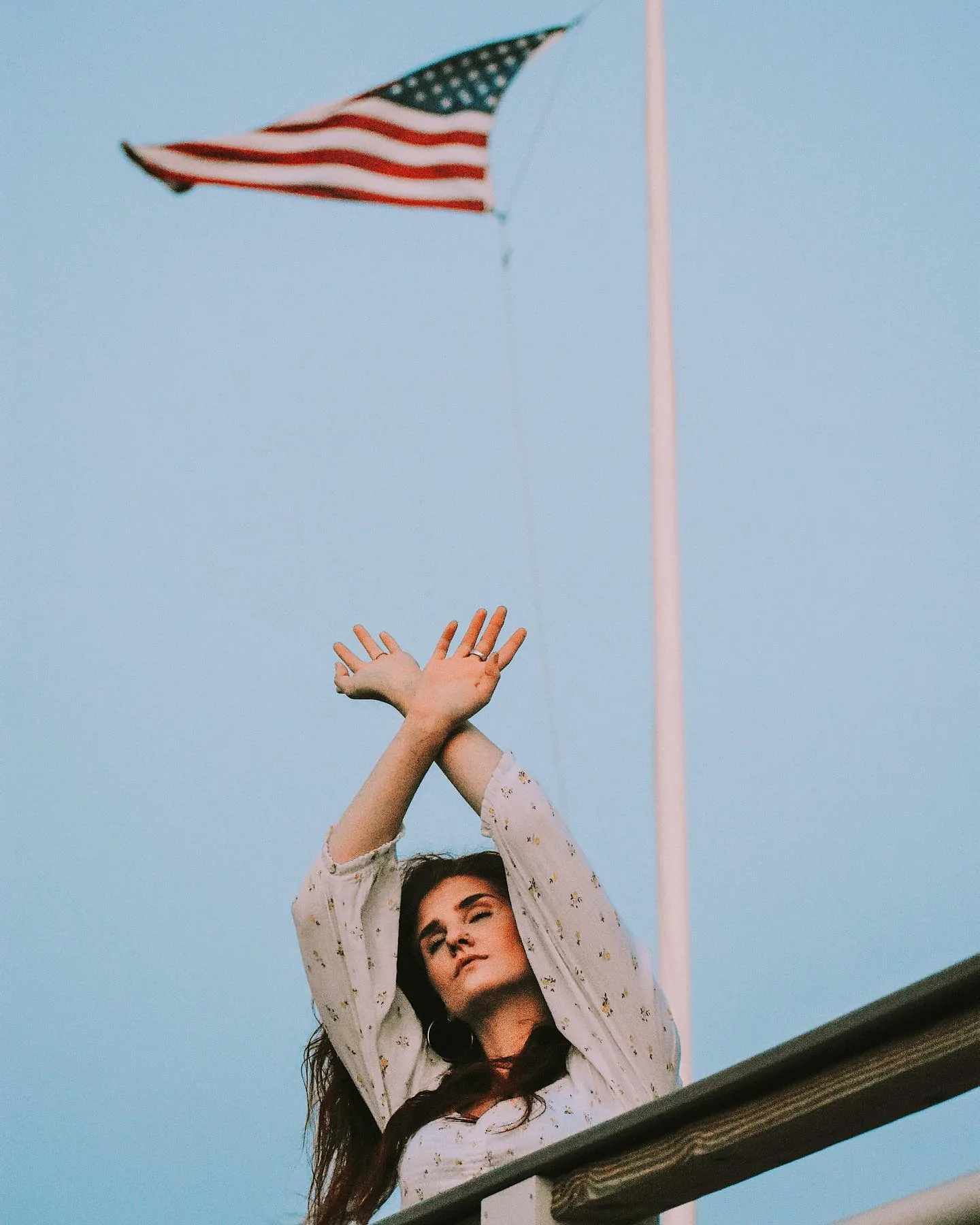 Woman in a white floral blouse leaning back over a railing with both arms crossed above her head, an American flag snapping from a pole in the pale blue sky above her.