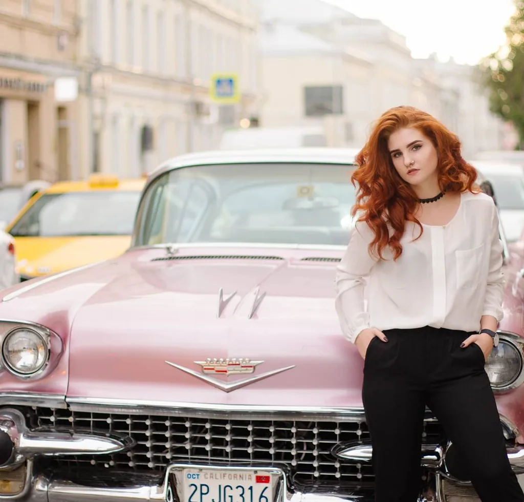 Daria in a white silk blouse and black trousers, red hair loose, leaning against the hood of a pink 1958 Cadillac with California plates on a Moscow street