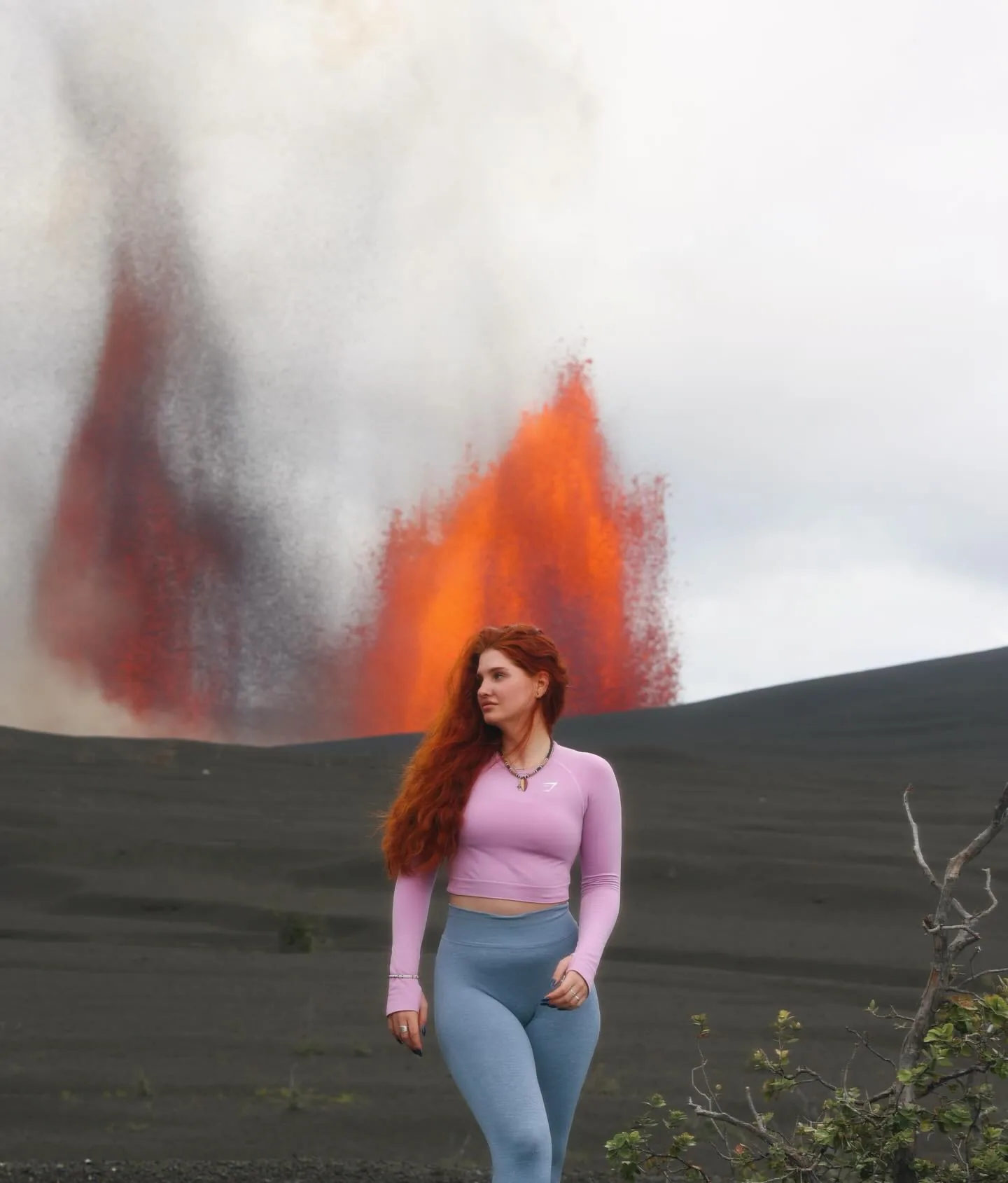 Daria in a pink long-sleeve crop top and grey leggings walking across a black cinder slope, looking off to one side, a tall orange lava fountain erupting behind her